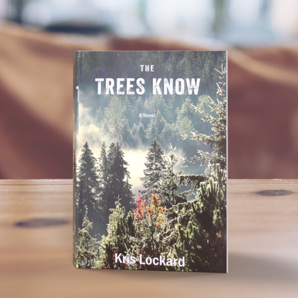 The Trees Know book sitting on a wooden table. The cover is of mist between mountainous trees